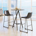 Two dark-colored bar stools with metal legs are placed around a round wooden table in a room with large windows.