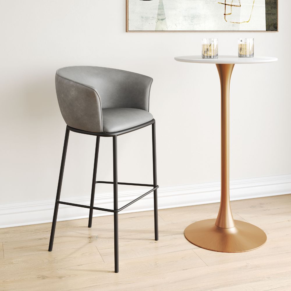 Gray bar stool next to a gold pedestal table in a room with a white wall and light wood floor.