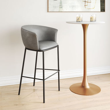 Gray bar stool next to a gold pedestal table in a room with a white wall and light wood floor.