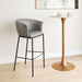 Gray bar stool next to a gold pedestal table in a room with a white wall and light wood floor.