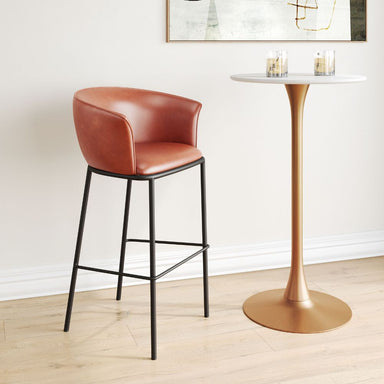 Brown leather bar stool next to a gold pedestal table with candles on a light wood floor.