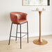 Brown leather bar stool next to a gold pedestal table with candles on a light wood floor.