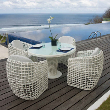 Outdoor dining set with a round table and four chairs on a wooden deck by a pool with ocean view.