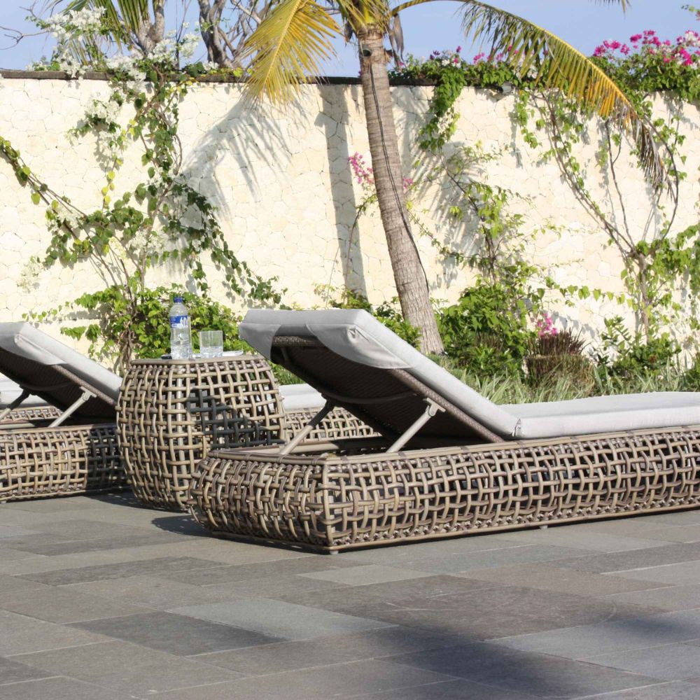 Wicker lounge chairs on a patio with a palm tree and flowers in the background