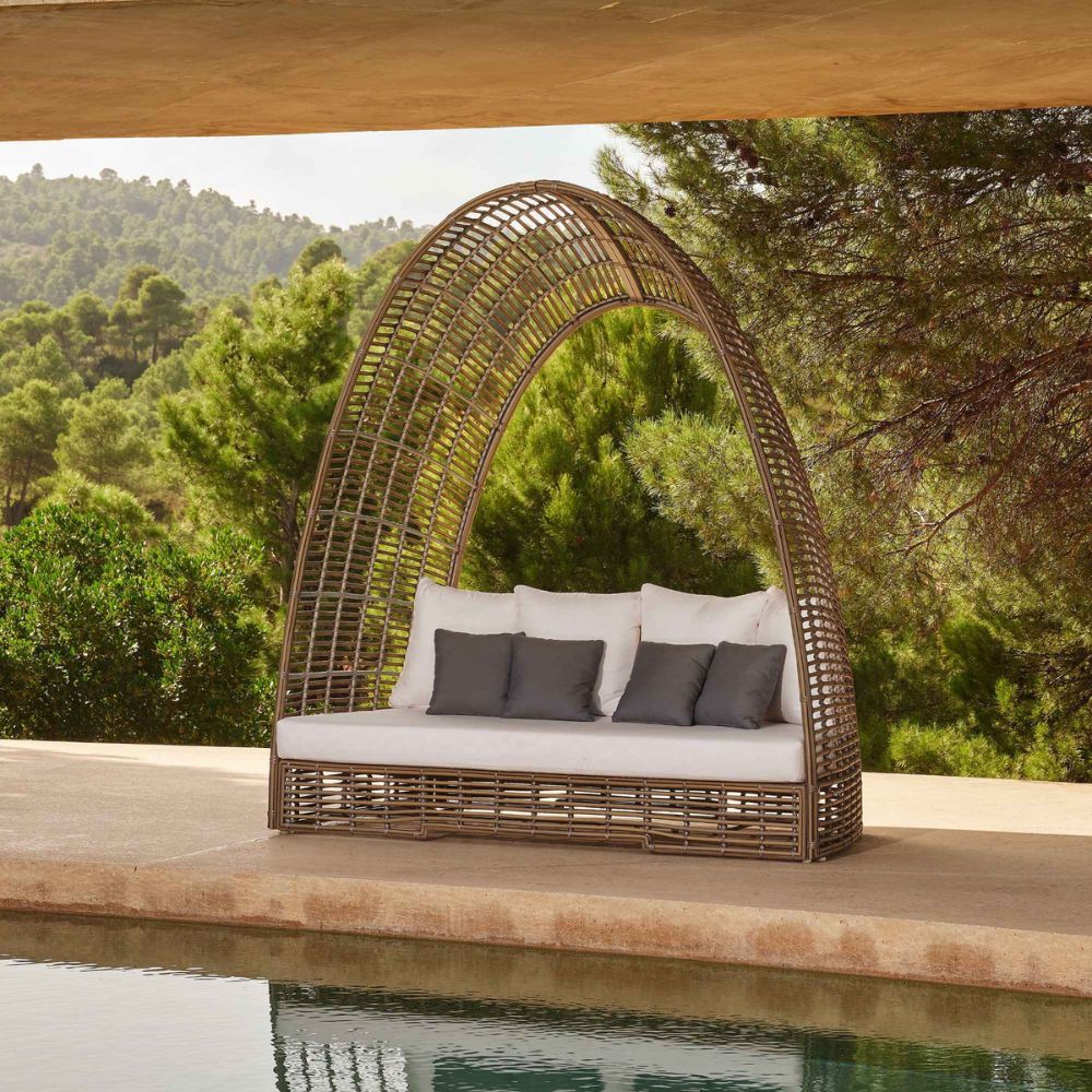 Wicker outdoor swing with white cushions and gray pillows by a poolside with greenery in the background