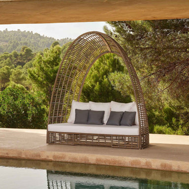 Wicker outdoor swing with white cushions and gray pillows by a poolside with greenery in the background