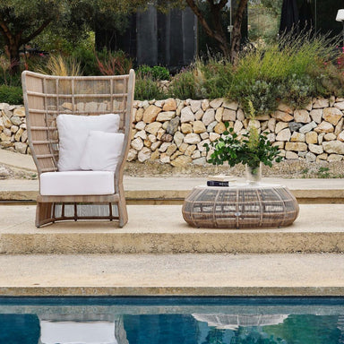 Wicker chair with white cushions and a matching ottoman by a poolside with a stone wall and garden in the background.