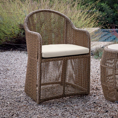 Wicker chair with a cushion on a gravel surface with plants in the background
