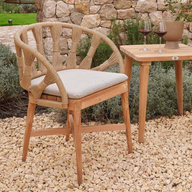 Wicker chair with a cushion on a pebble-covered ground, next to a wooden table with glasses of red wine.