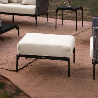 White cushioned ottoman on a brown outdoor rug with a blurred background of outdoor furniture.