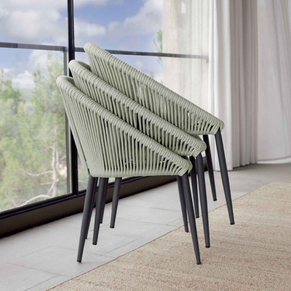 Three green woven chairs with black legs arranged near a large window with a view of trees.