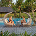 Two women sitting in wicker chairs by a pool with a tropical setting.