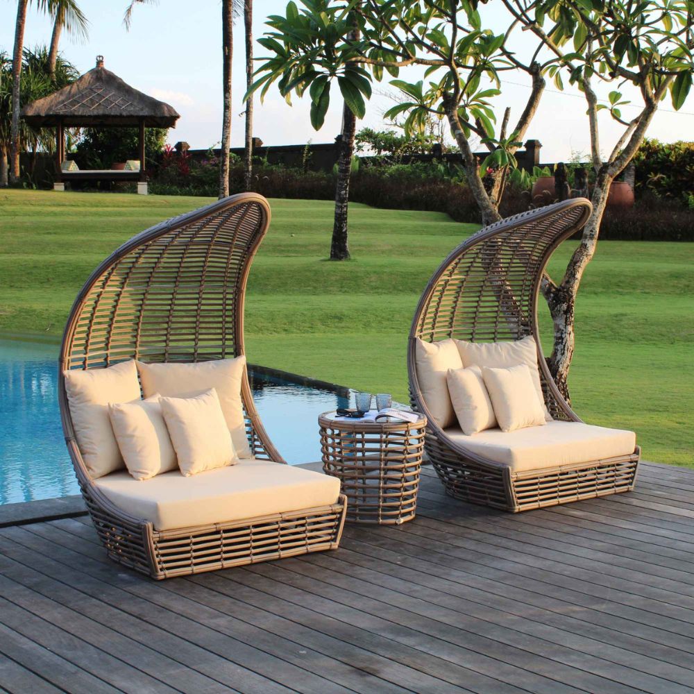 Two wicker chairs with cushions on a wooden deck by a poolside, surrounded by greenery.