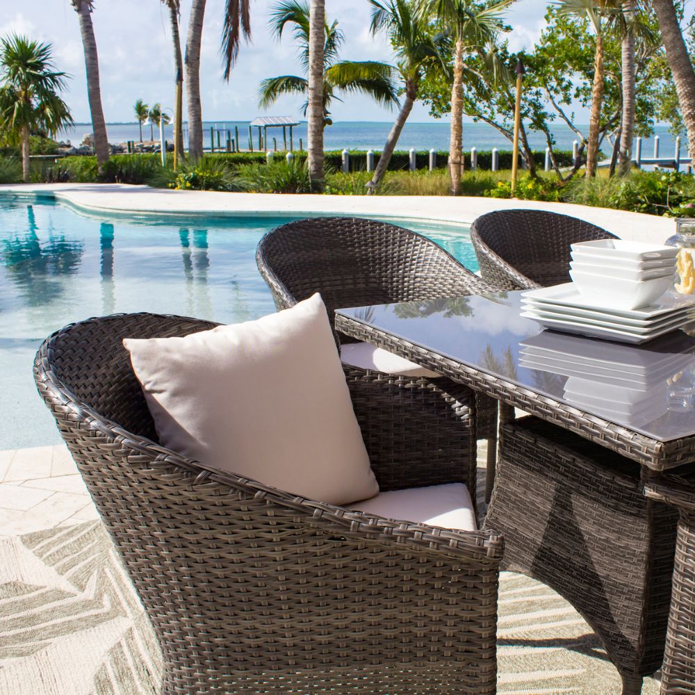 Outdoor patio set with wicker chairs and table by a poolside with palm trees and ocean view.