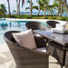Outdoor patio set with wicker chairs and table by a poolside with palm trees and ocean view.