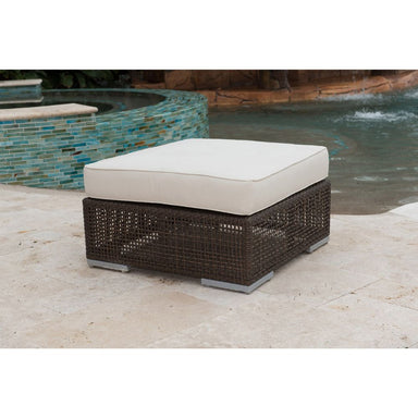 Wicker ottoman with a white cushion on a patio near a pool.