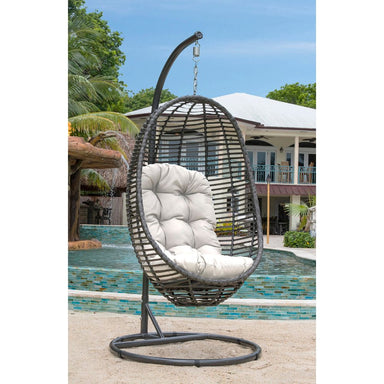 Hanging egg-shaped chair with white cushion in front of a pool and house.
