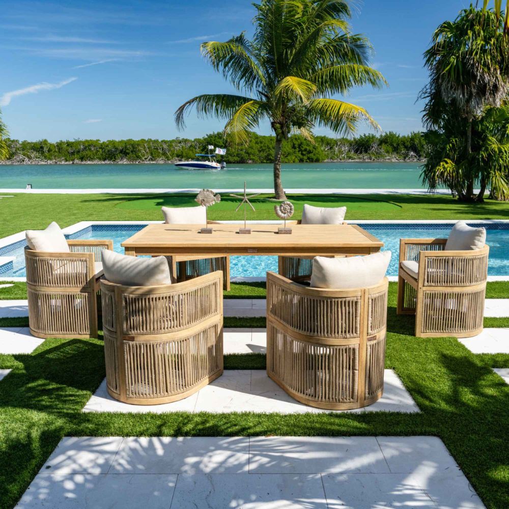 Outdoor dining set with wooden chairs and table by a poolside with palm trees and water in the background.