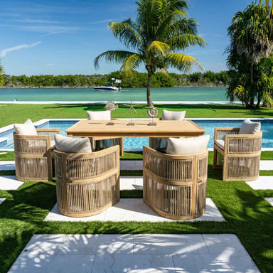 Outdoor dining set with wooden chairs and table by a poolside with palm trees and water in the background.