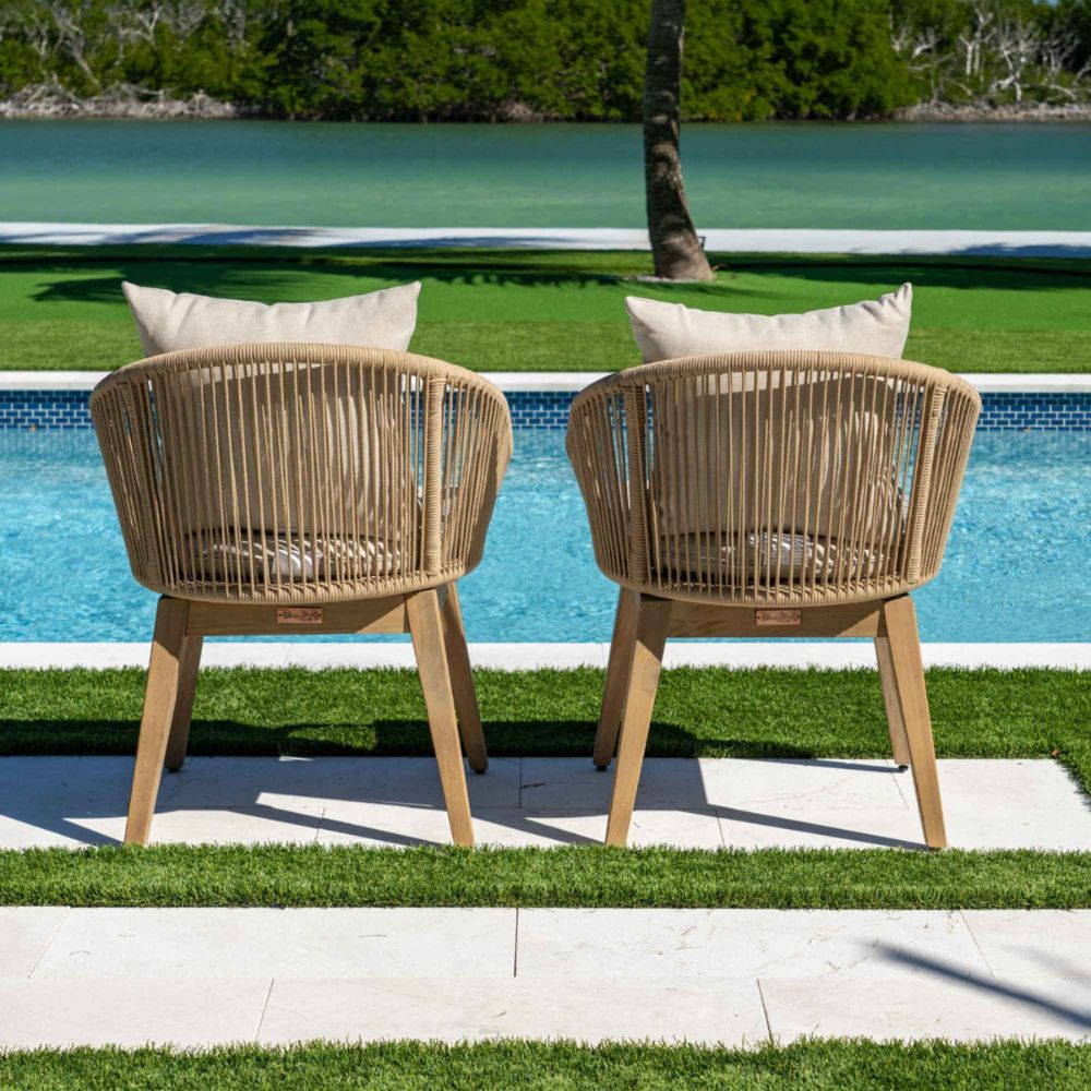 Two wicker chairs with cushions by a poolside with greenery in the background