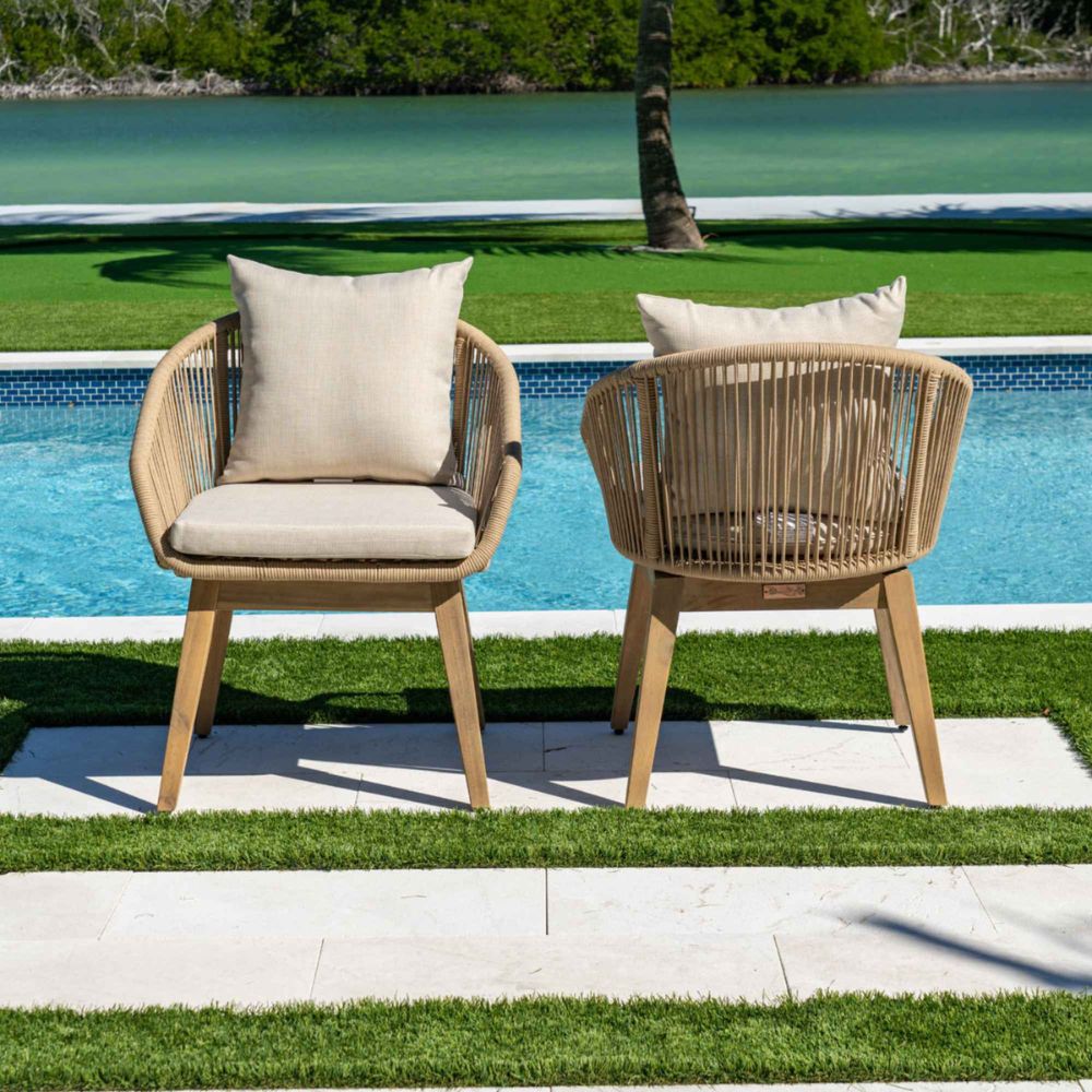 Two wicker chairs with beige cushions by a poolside with greenery in the background