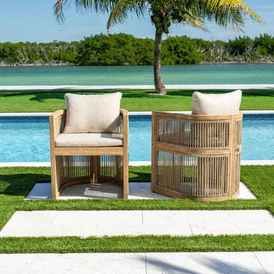 Two wooden outdoor chairs with white cushions by a poolside with a palm tree in the background.