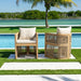 Two wooden outdoor chairs with white cushions by a poolside with a palm tree in the background.