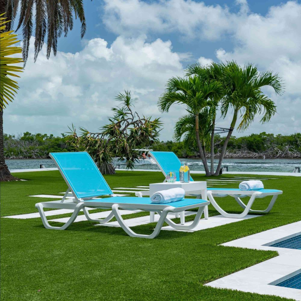 Two blue lounge chairs on a grassy area near a pool with palm trees and water in the background.
