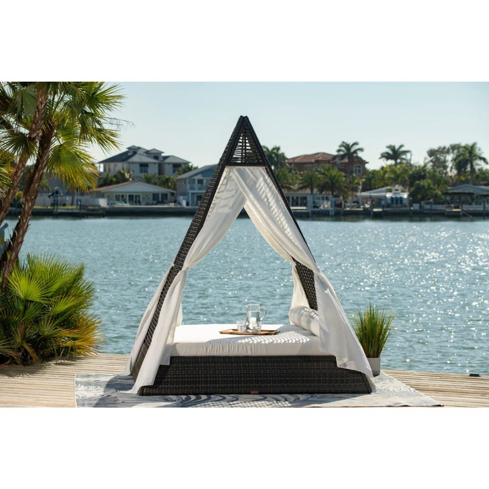 Outdoor canopy bed with mosquito net on a dock by a body of water with houses and palm trees in the background.