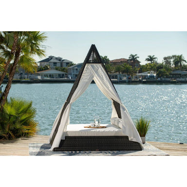 Outdoor canopy bed with mosquito net on a dock by a body of water with houses and palm trees in the background.