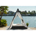 Outdoor canopy bed with mosquito net on a dock by a body of water with houses and palm trees in the background.