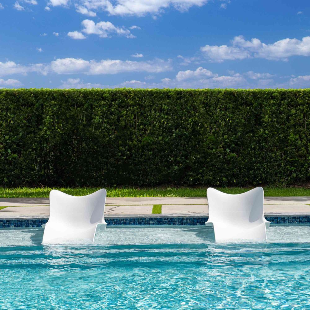 Two white chairs in a pool with a hedge and blue sky in the background