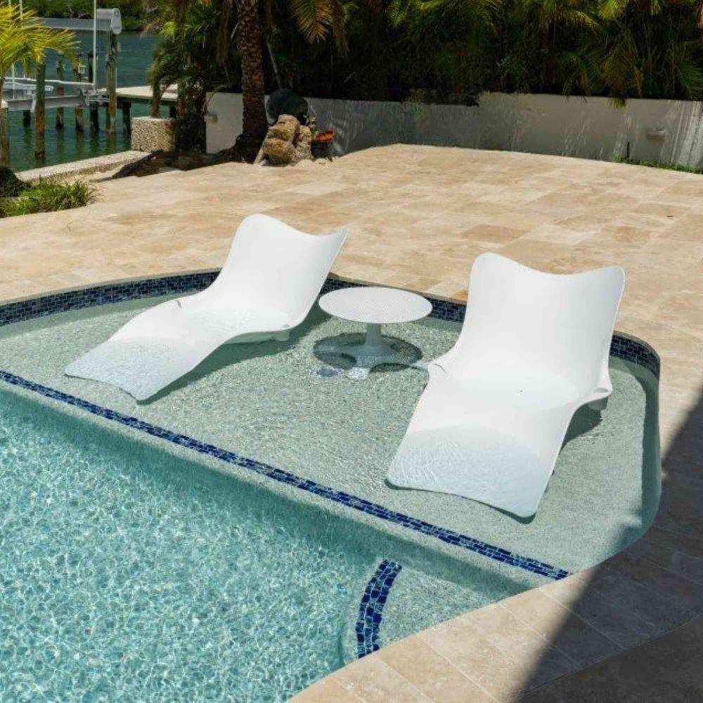 Two white lounge chairs and a small table by a pool with a waterfront view.