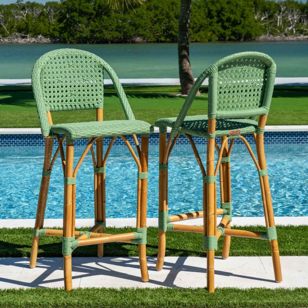 Two green and brown wicker bar stools by a poolside with a scenic background.