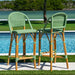 Two green and brown wicker bar stools by a poolside with a scenic background.