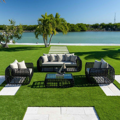 Outdoor patio set with black wicker furniture and white cushions on a grassy area by a waterfront.