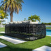 Black woven outdoor sofa with cushions on a grassy area with palm trees and a pool in the background.