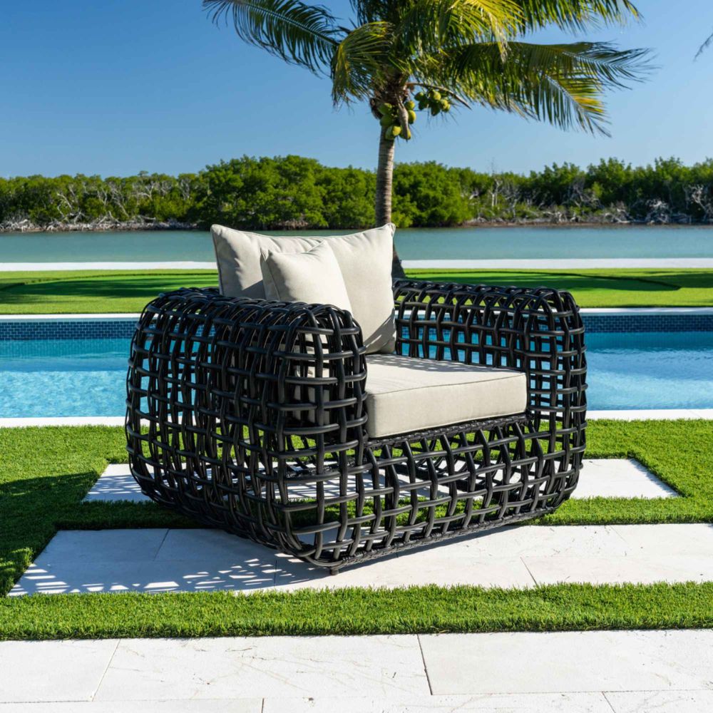 Black wicker chair with white cushions on a patio by a poolside with palm trees in the background.
