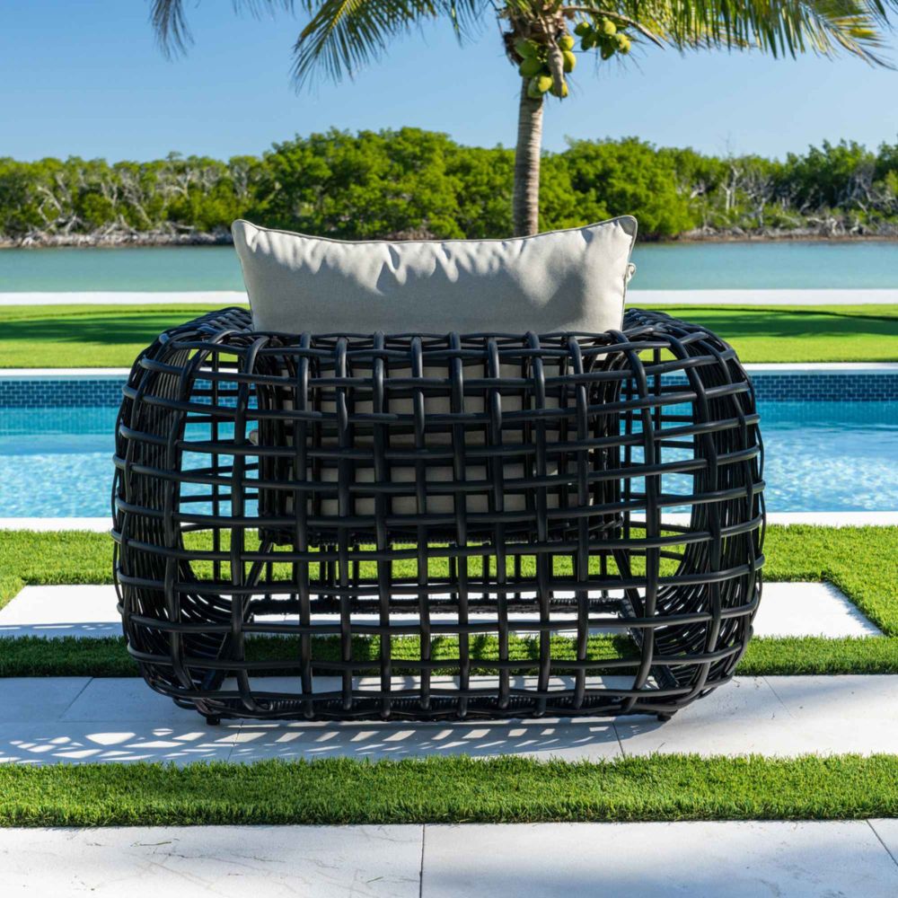 Black woven outdoor chair with a white cushion on a patio with a pool and greenery in the background.