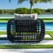 Black woven outdoor chair with a white cushion on a patio with a pool and greenery in the background.